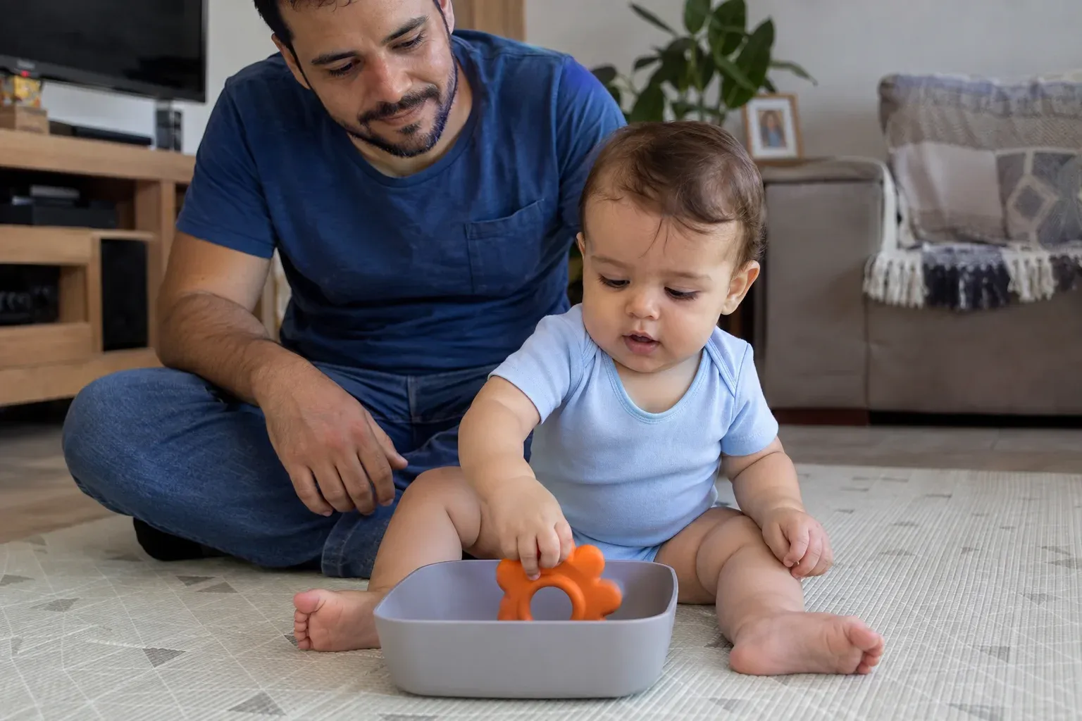 Bebê retirando um brinquedo de um recipiente baixo e aberto, com o pai ao lado.